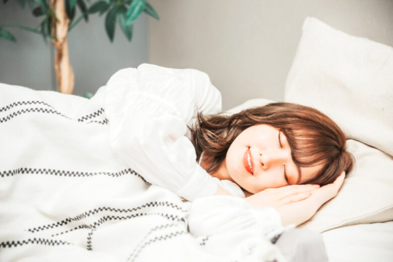 白い長袖を着た人がベッドで眠っている様子。白い布団と観葉植物が映る、落ち着いた夜の室内。
