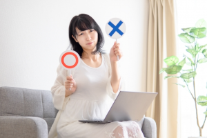 ソファに座っている女性がひざにパソコンを乗せながら〇✕のプレートを持っている