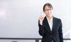 ホワイトボードの前で赤い眼鏡をかけた女性が人差し指を立てている