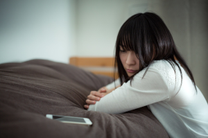 ベッドに置いたスマホを暗い表情で眺めている女性