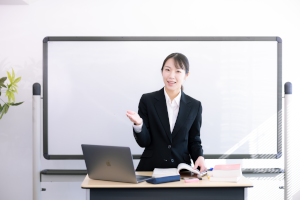 ホワイトボードをバックに、スーツを着た女性が「どうぞ」と言っている様子
