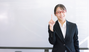 ホワイトボードをバックに、赤い眼鏡をかけたスーツの女性が、人差し指を立てている