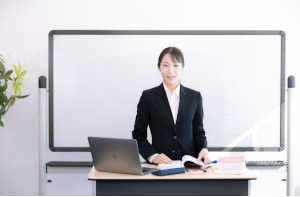 ホワイトボードの前にスーツを着た女性がいて、教卓にパソコンや辞書などが置いてある