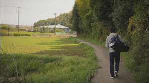田んぼと林の間を歩く大きなカバンを持った男性の後ろ姿