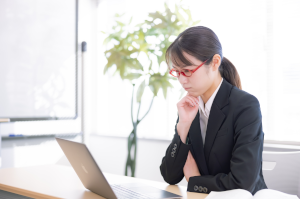 パソコンの前で顎に手をあてて考え事をしている赤い眼鏡をかけたスーツの女性