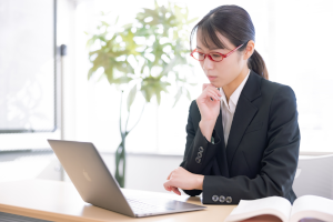 パソコンを前に座りながら考え事をしているスーツの女性