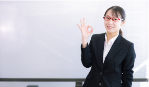 ホワイトボードをバックに、OKと指で作っている赤い眼鏡をかけたスーツの女性