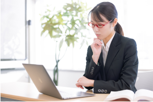 パソコンの前で考え事をしている赤い眼鏡をかけたスーツの女性