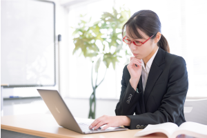 赤い眼鏡をかけたスーツの女性がパソコンを見て悩んでいる様子