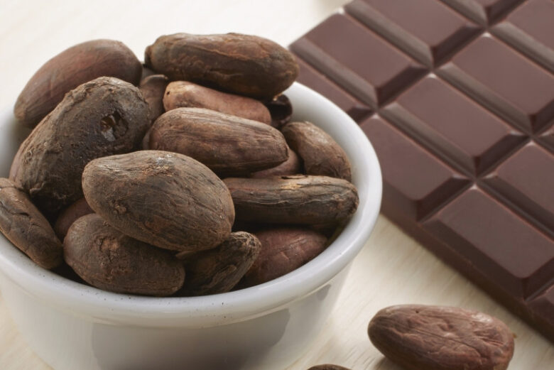 白い器に入ったカカオ豆と、その横に並べらた光沢のあるダークチョコレートバー。木目のテーブルの上にカカオ豆が散らばっている。