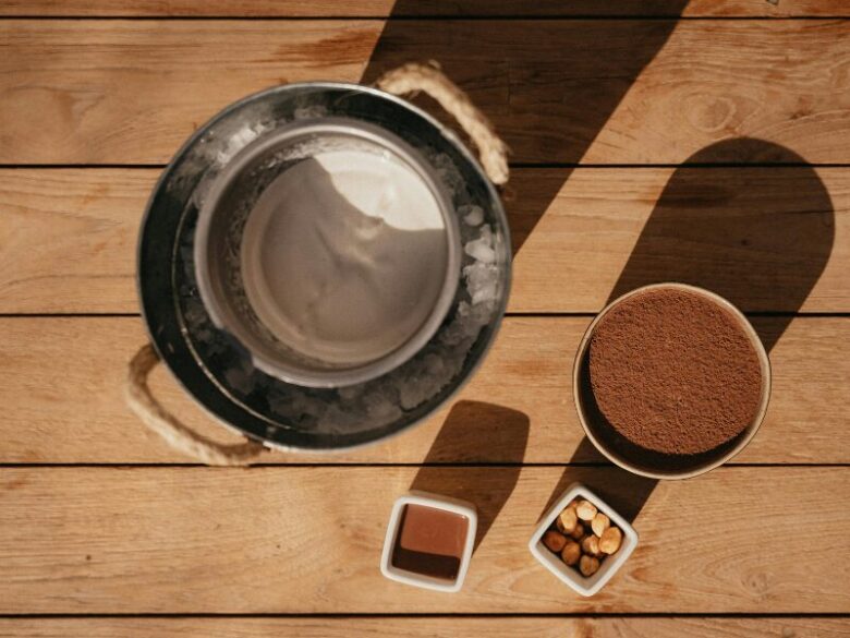 氷を入れたバケツにクリーム状の白い食材が入った器を乗せ、隣にココアパウダー、チョコレートソース、ヘーゼルナッツを並べた木製のテーブルの上のデザート準備シーン。
