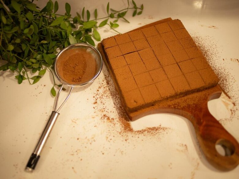 ココアパウダーがまぶされたチョコレートの四角いブロックが木のカッティングボードに整然と並び、横には茶こしと緑のハーブが添えられている。