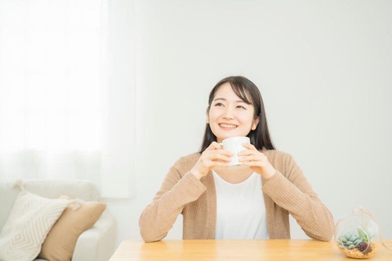 白いマグカップを両手で持ち、リラックスした表情で座っている人物。木製テーブルの上にはガラスのテラリウムが置かれ、背景には柔らかな光が差し込むカーテンとソファが見える。