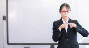 赤い眼鏡をかけたスーツの女性が指で小さく×を作っている