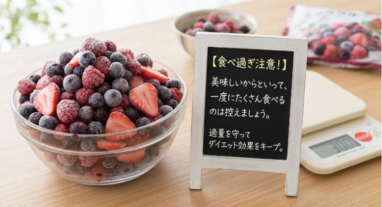 木製のテーブルの上に、山盛りにされた冷凍フルーツが入ったガラスのボウルと、隣には「【食べ過ぎ注意！】美味しいからといって、一度にたくさん食べるのは控えましょう。適量を守ってダイエット効果をキープ。」と書かれたミニ黒板風の看板が置かれている。背後にはデジタルスケール（キッチンスケール）やパッケージも写っている。