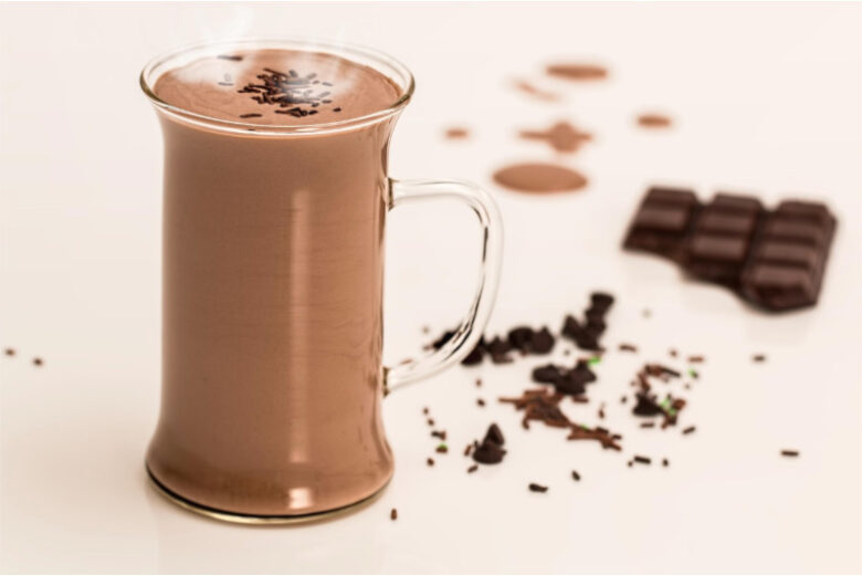 チョコレートスプレーがトッピングされたホットチョコレートの入ったガラスマグカップ。湯気が立ちのぼり、周囲にはちょこれーとのかけらやココアパウダーが散りばめられている。