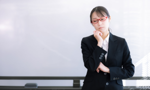 赤い眼鏡をかけたスーツの女性が顎に手をあてて悩んでいる