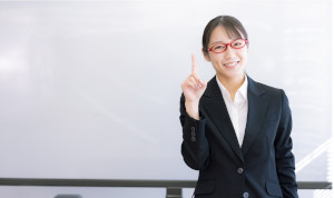 赤い眼鏡をかけたスーツの女性が人差し指を立てている