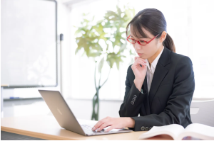 赤い眼鏡をかけたスーツの女性がパソコンの前で考え事をしている