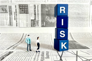 敷かれた英字新聞の上にRISKと書かれた縦に積まれた積み木4つとおもちゃの人形