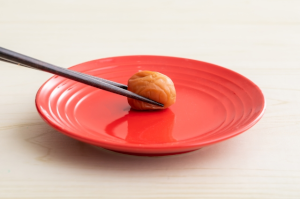 赤い丸皿に乗った1個の梅干しを箸で取ろうとしている所
