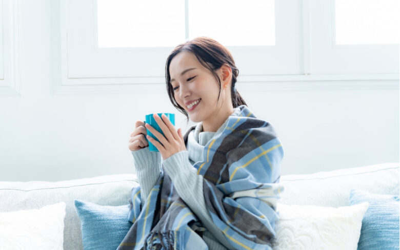 ブランケットに包まれてソファで温かい飲み物を持つ女性の手元の。