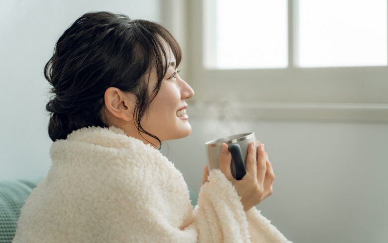 ブランケットにくるまった妊婦さんが、湯気の立つホットココアを両手で包んで飲んでいる様子
