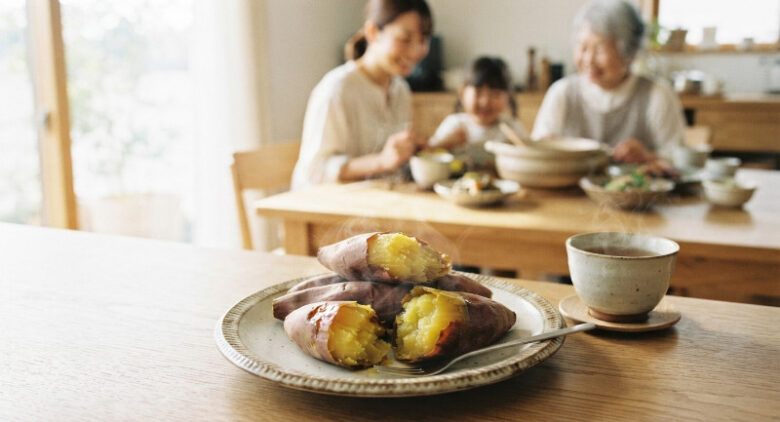 湯気が立つホクホクの焼き芋が盛られたお皿が手前に置かれ、奥のダイニングテーブルでは家族が笑顔で食事を楽しんでいる温かい雰囲気の様子。