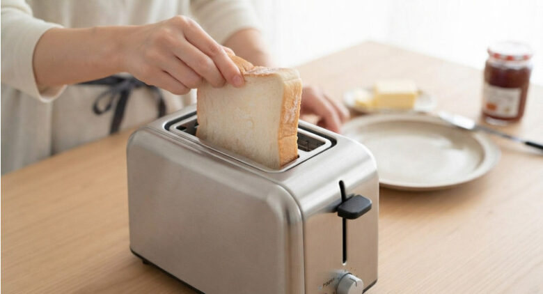 清潔な木製のキッチンの上で、凍ったままの厚切り食パンをステンレス製トースターのスロットに手で入れている様子。