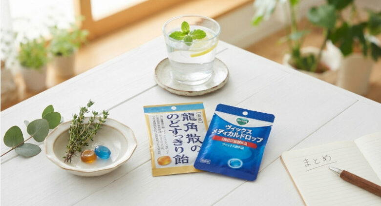 白い木製のテーブルの上に、龍角散のどすっきり飴とヴィックス メディカルドロップのパッケージ、レモンとミントが入った水のグラス、そして「まとめ」と書かれたノートがきれいに配置されている様子。