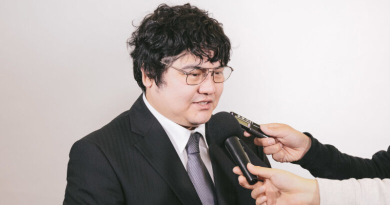 記者会見で多くのカメラやマイクに囲まれ、真剣な表情でインタビューに応じるスーツ姿の男性